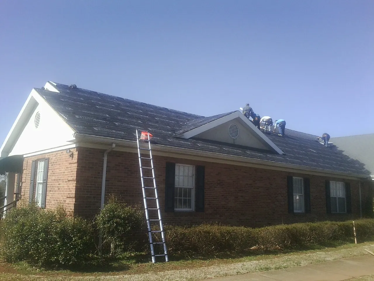 Crew tearing shingles off a brick ranch roof with a ladder set — Metal Roof Repair work by our crew in Bazine