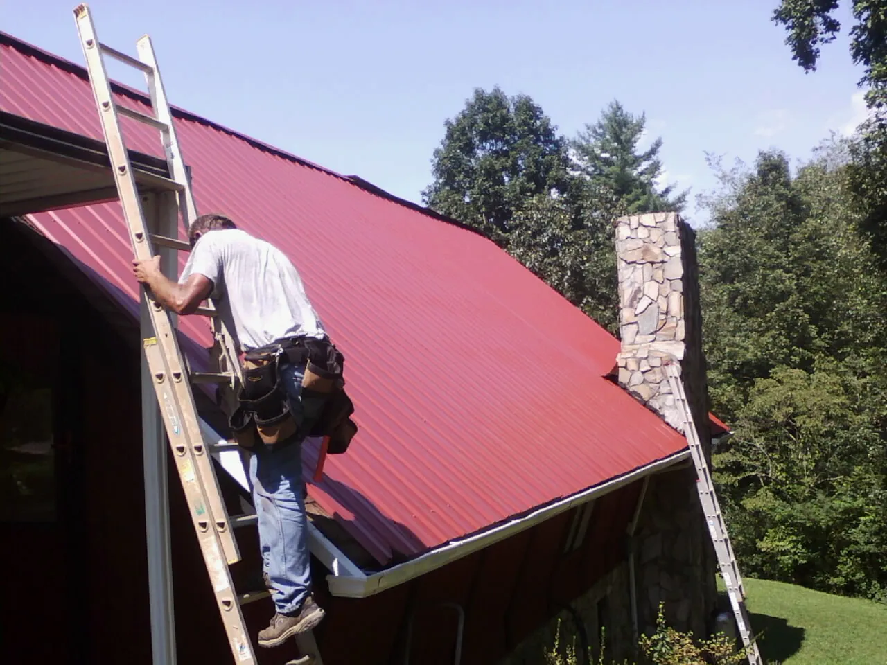 Roofer on a ladder beside a red metal roof and stone chimney — Soffit & Fascia Repair by Roofing Brothers in Bazine